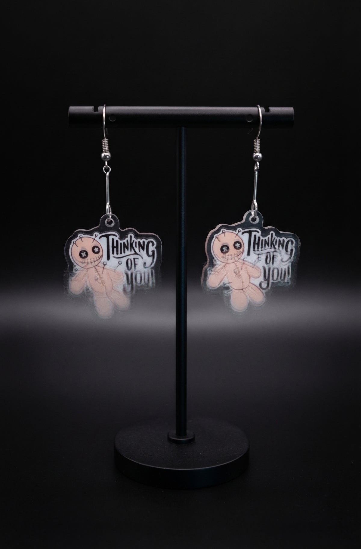 Voodoo Doll Thinking of You Earrings by Circle of Heather, printed acrylic voodoo doll charms with pins and stitch detail on silver-tone bar connectors, hanging on a black T-bar jewelry stand.