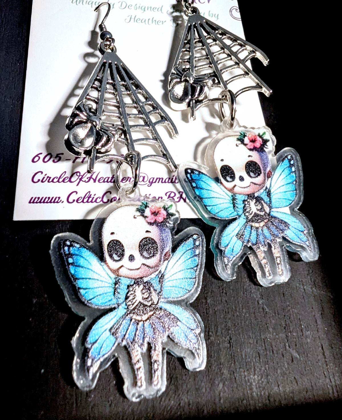 Webbed Skull Fairy Earrings by Circle of Heather worn by a model with a dark blue and purple pixie cut, silver-tone spider web charm above a printed skull fairy with blue butterfly wings, against a dark studio background.