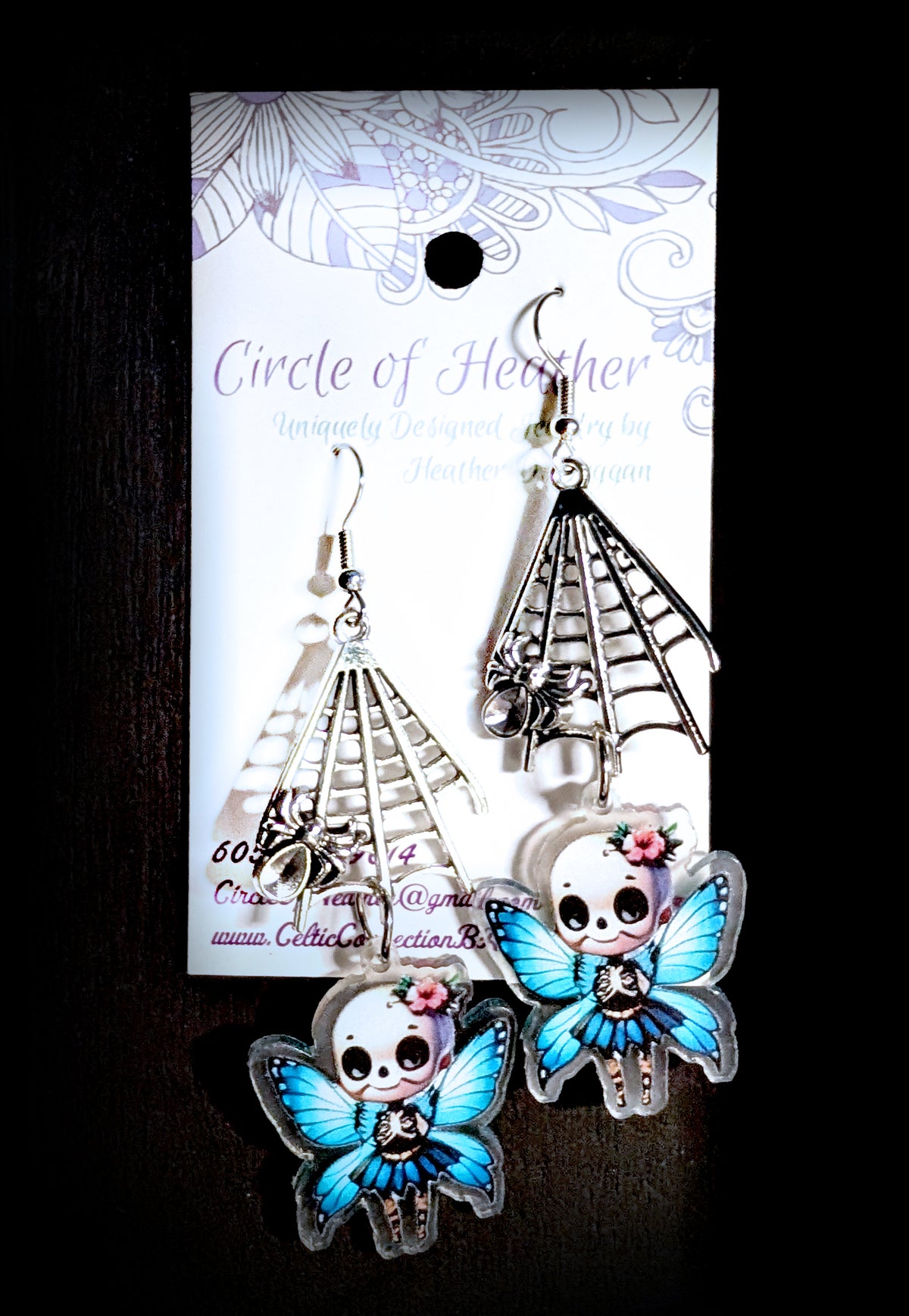 Webbed Skull Fairy Earrings by Circle of Heather, full view of both earrings showing the silver-tone spider web charms with spider detail and printed acrylic skull fairy charms with blue butterfly wings and blue tutus, on a plain black background.