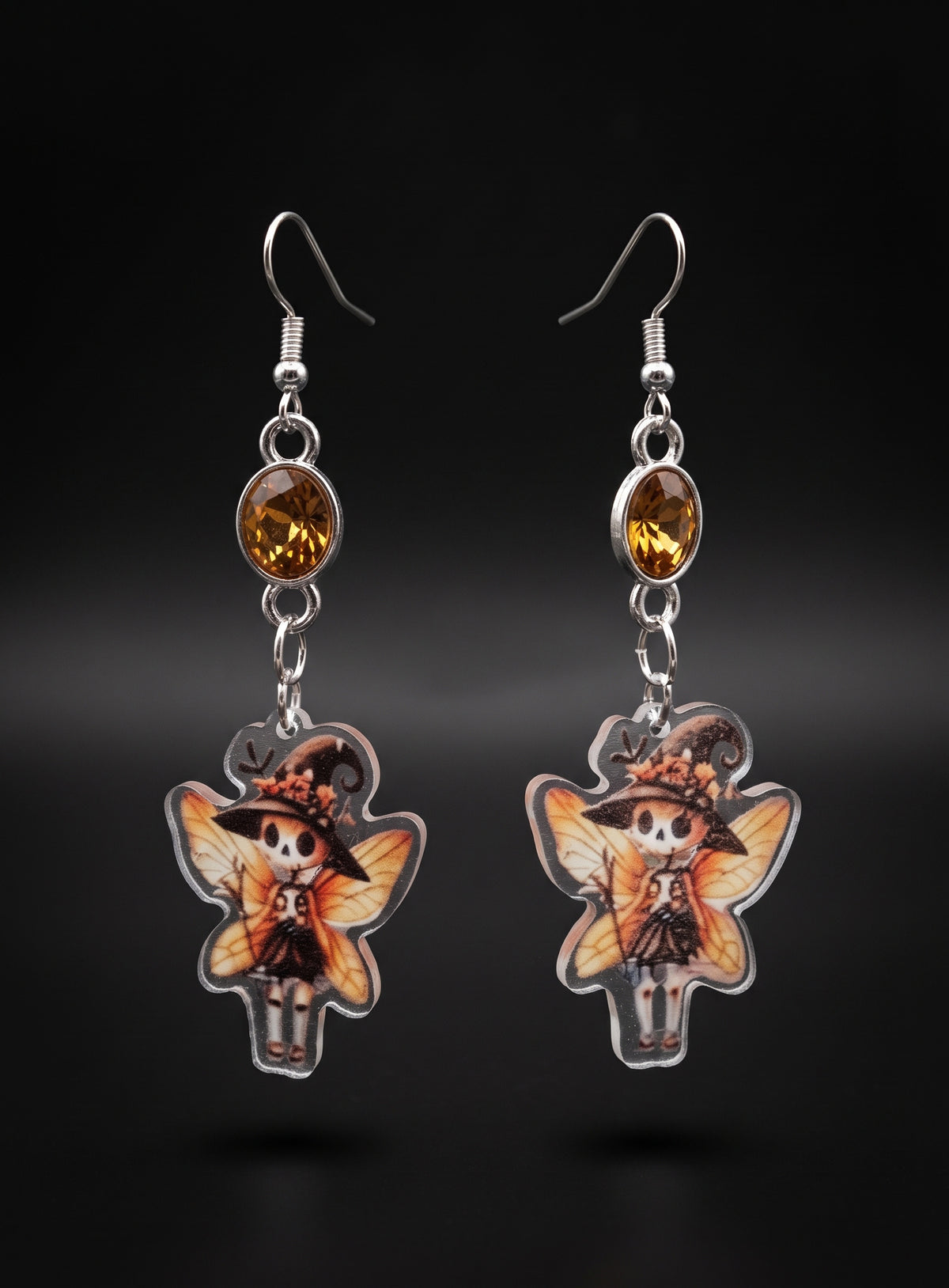 Witchy Skull Fairy Earrings by Circle of Heather, full view of both earrings showing the amber stone tops and skull fairy charms with flower-trimmed witch hats, orange wings and brooms, on a plain black background.