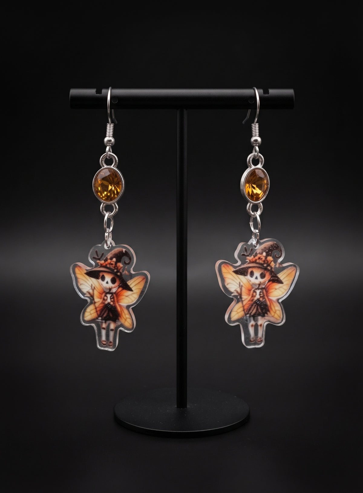 Witchy Skull Fairy Earrings by Circle of Heather, faceted amber stones in silver-tone bezels above printed acrylic skull fairy charms with witch hats and orange wings, hanging on a black T-bar jewelry stand.