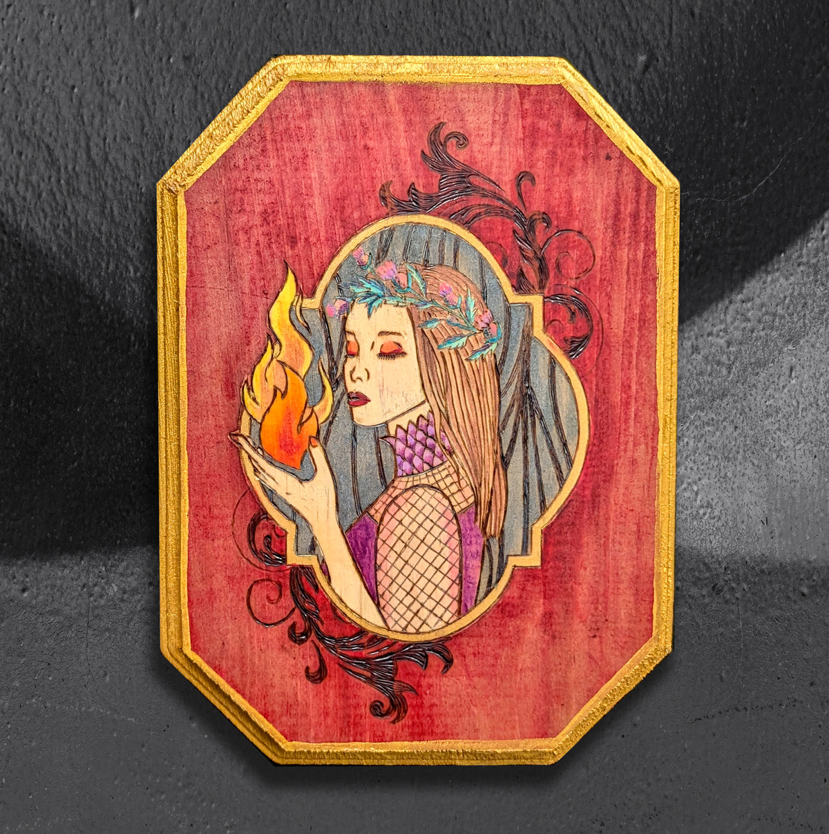 Hestia's Hearth by CamiThunderInk, an original pyrography and acrylic painting on a wood canvas showing the goddess Hestia holding a steady flame, framed by wood burned filigree and a metallic gold beveled edge