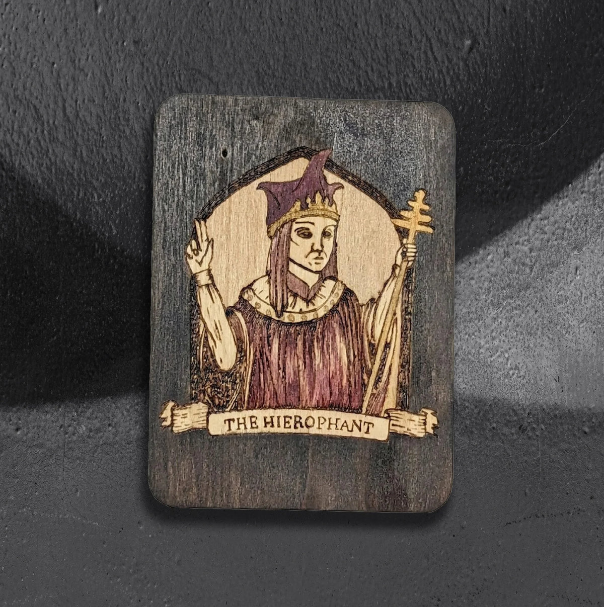 The Hierophant by CamiThunderInk, an original pyrography and watercolor tarot card on a thin wood panel, showing a crowned figure in purple robes raising one hand and holding a triple cross staff, framed by an arched halo with a banner reading The Hierophant, photographed on a gray textured background