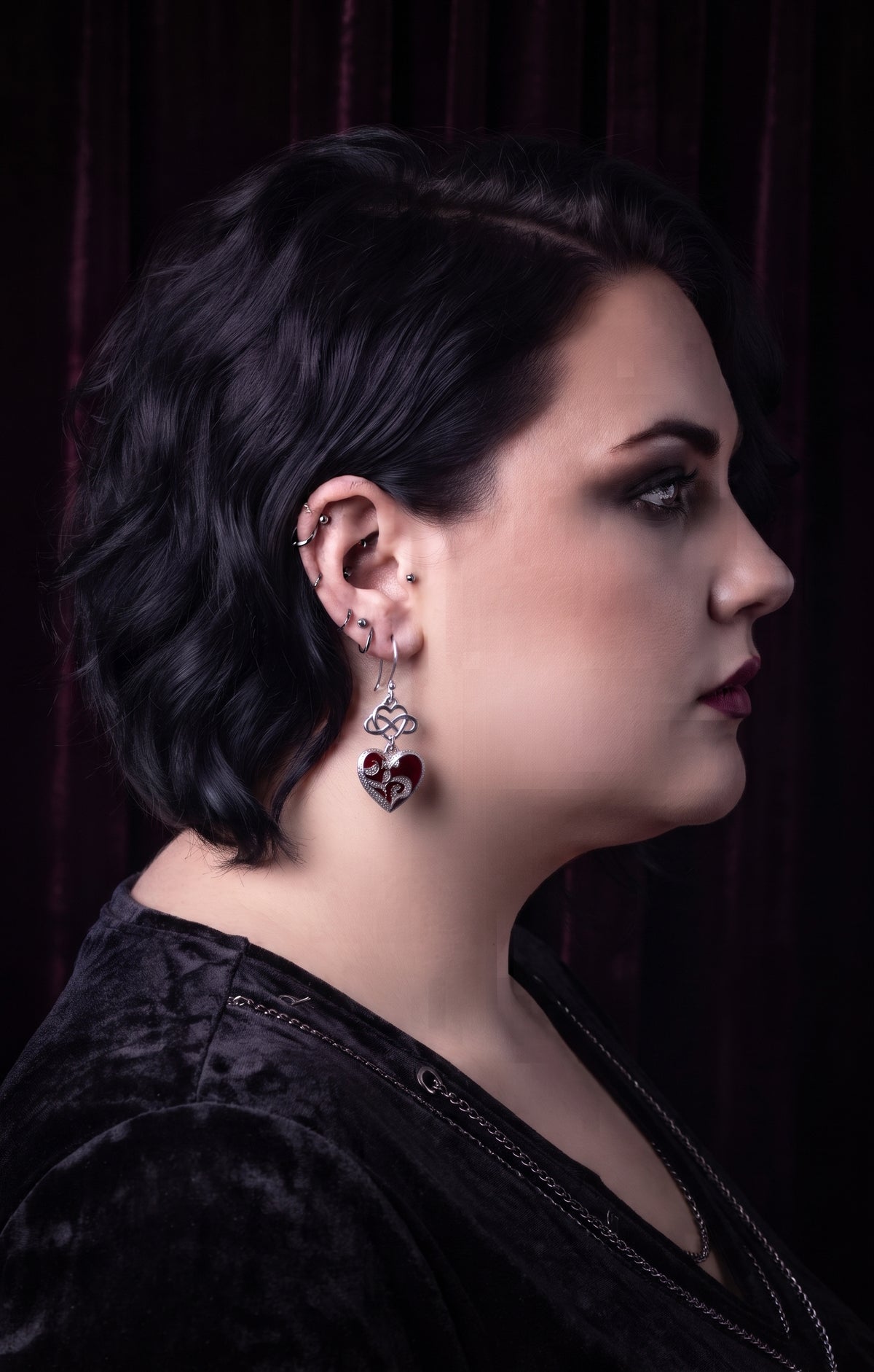 Model shown in profile wearing Infinity Hearts Earrings by Circle of Heather, silver-tone infinity heart knot above a filigree heart in deep red enamel