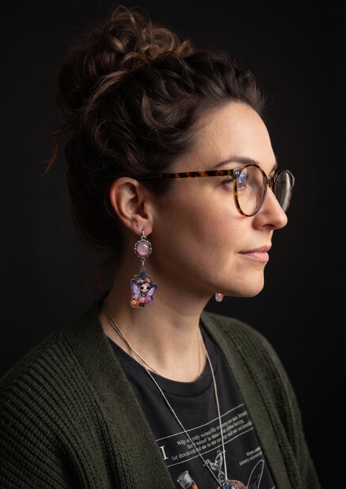 Treat Skull Fairy Earrings by Circle of Heather worn by a model with a curly dark updo and tortoiseshell glasses, pink cabochon above a printed skull fairy charm with a witch hat and pink wings, shown with a green cardigan.