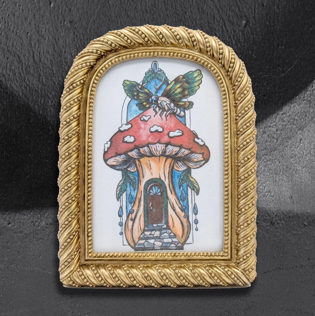 Mushie House framed print by CamiThunderInk, a whimsical mushroom cottage illustration topped with a winged insect, in a small arched gold frame with rope and bead trim