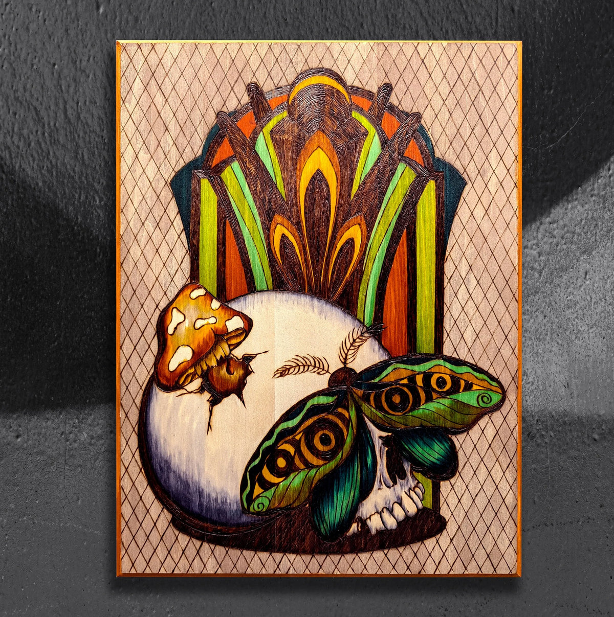 Another view of MushMoth Skull by CamiThunderInk, an original pyrography, watercolor, and acrylic painting on wood showing a human skull with a mushroom growing through a crack and an emerald green moth resting over the eyes, in front of a burned Art Deco lattice screen