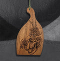 Mycelium Catworks cutting board by CamiThunderInk, an acacia wood cutting board with a hand burned drawing of a cat skull overgrown with flowers and leafy stems, hanging from a sage green cord against a gray textured background