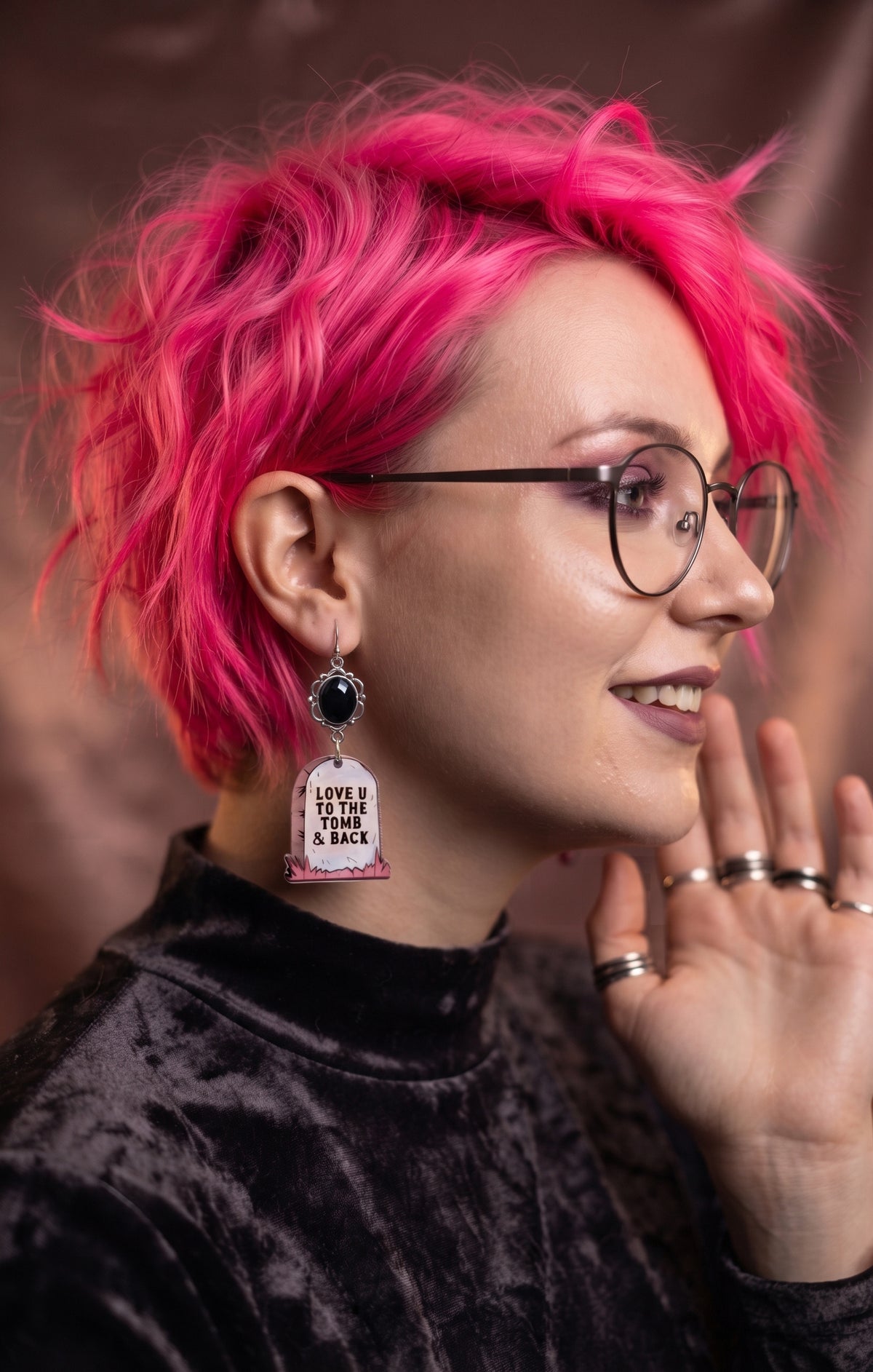 Love U to the Tomb and Back Earrings by Circle of Heather worn by a model with bright pink hair and wire glasses, faceted black stone above a printed headstone charm reading "Love U to the tomb and back," shown with a black crushed velvet top.