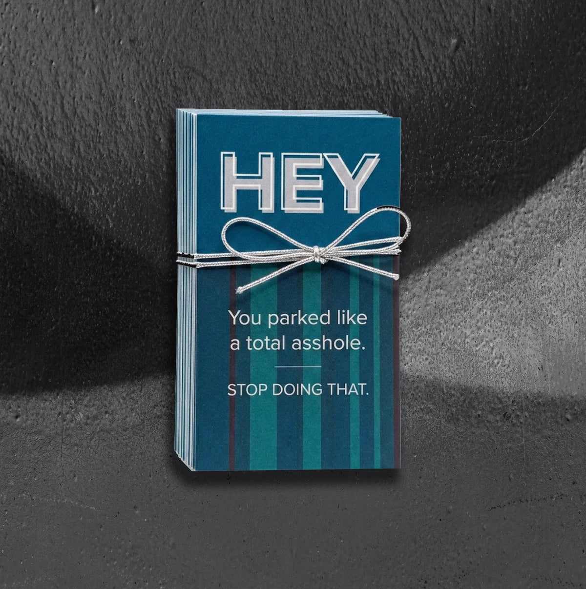 A stack of blue striped parking cards by Reclamation Tattoo and Oddities, tied with a silver string bow, with the top card reading "HEY" in large letters and "You parked like a total asshole. STOP DOING THAT." below, photographed on a gray textured background