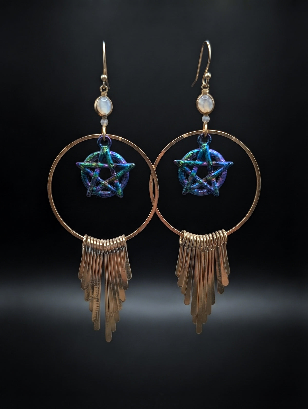 A pair of bohemian-style gold-tone hoop earrings with a central iridescent blue and purple pentagram star, a small round white stone, and a dangling fringe of thin gold-tone bars, set against a dark background.
