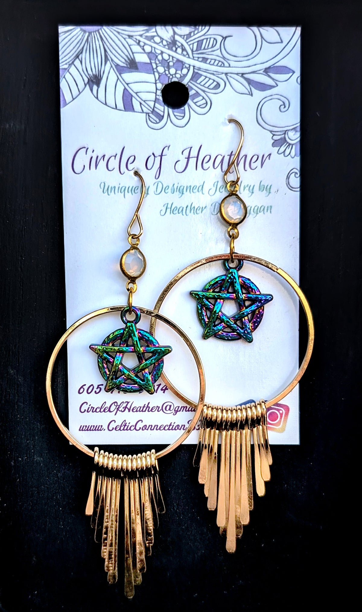 A pair of gold-toned dangle earrings displayed on a branded card. Each earring has a large circular hoop, a pentagram star charm with an iridescent blue-green finish, and a fringe of small gold pins hanging below.