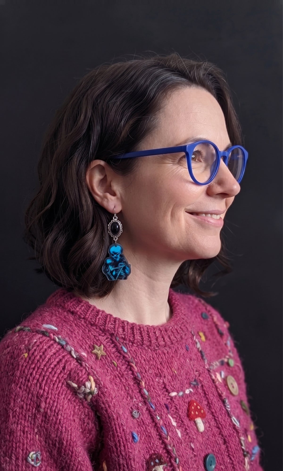 Skele Kitty Snuggles Earrings by Circle of Heather worn by a model with a dark wavy bob and blue glasses, black cabochon above a blue and black printed charm of a skeleton holding a cat, shown with a pink embroidered sweater.