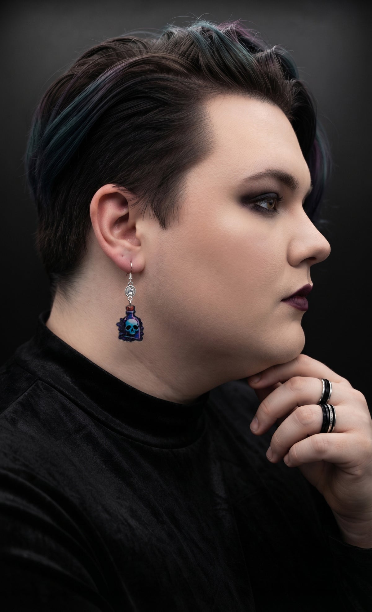 Skull Potion Earrings by Circle of Heather worn by a model with dark hair and teal streaks, clear rhinestone above a printed blue and purple potion bottle charm with a skull inside, against a dark studio background.