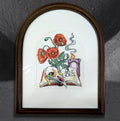So It Goes by CamiThunderInk, an original colored pencil drawing of a raven skull, a candle, and poppies on an open book with scattered gummy worms, in an arched frame