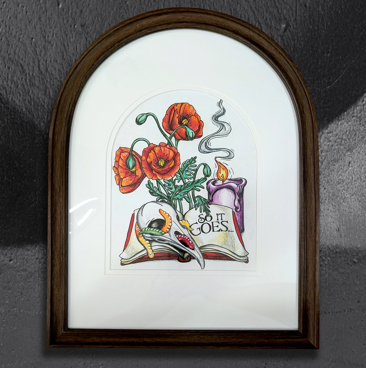 So It Goes by CamiThunderInk, an original colored pencil drawing of a raven skull, a candle, and poppies on an open book with scattered gummy worms, in an arched frame