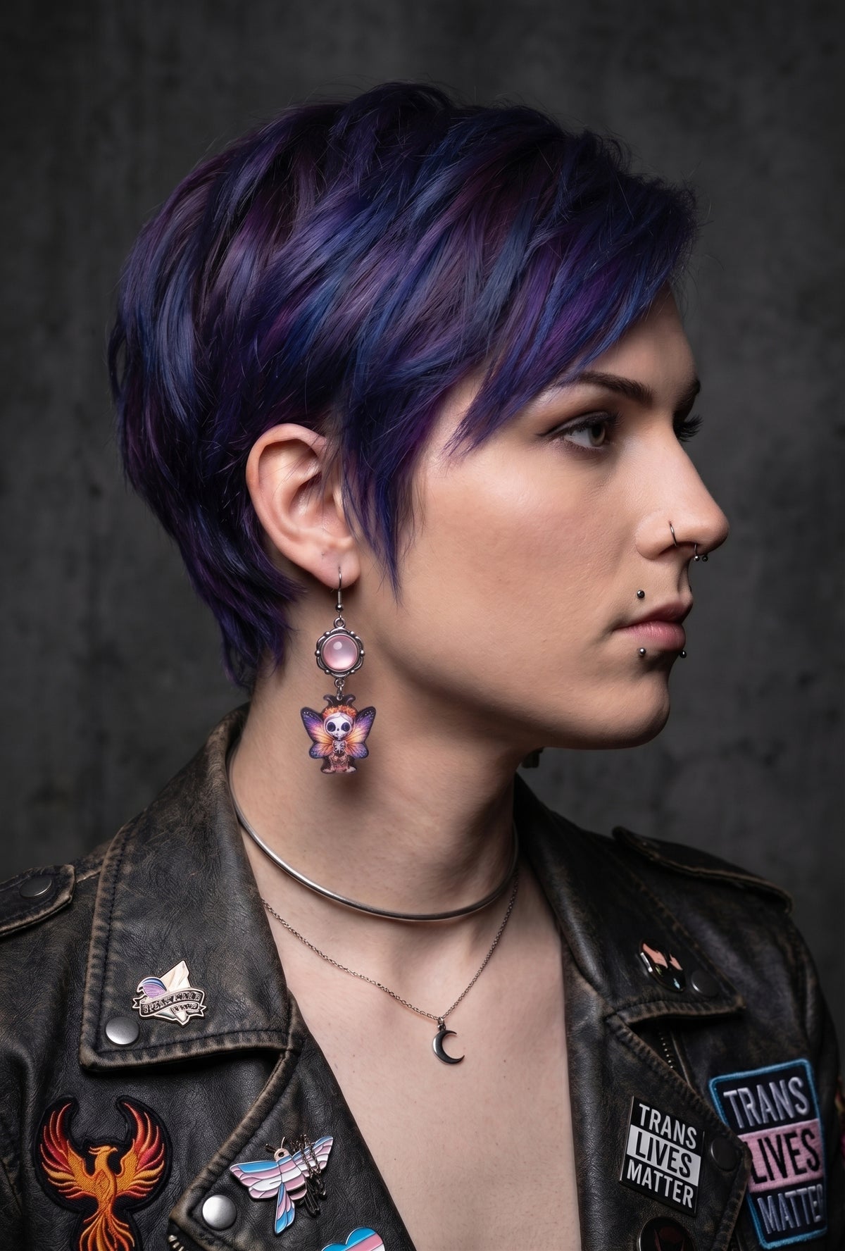 Sweet Pink Skull Fairy Earrings by Circle of Heather worn by a model with a purple and blue pixie cut, pale pink cabochon above a printed skull fairy charm with pink and orange butterfly wings, shown with a patched leather jacket against a gray wall.