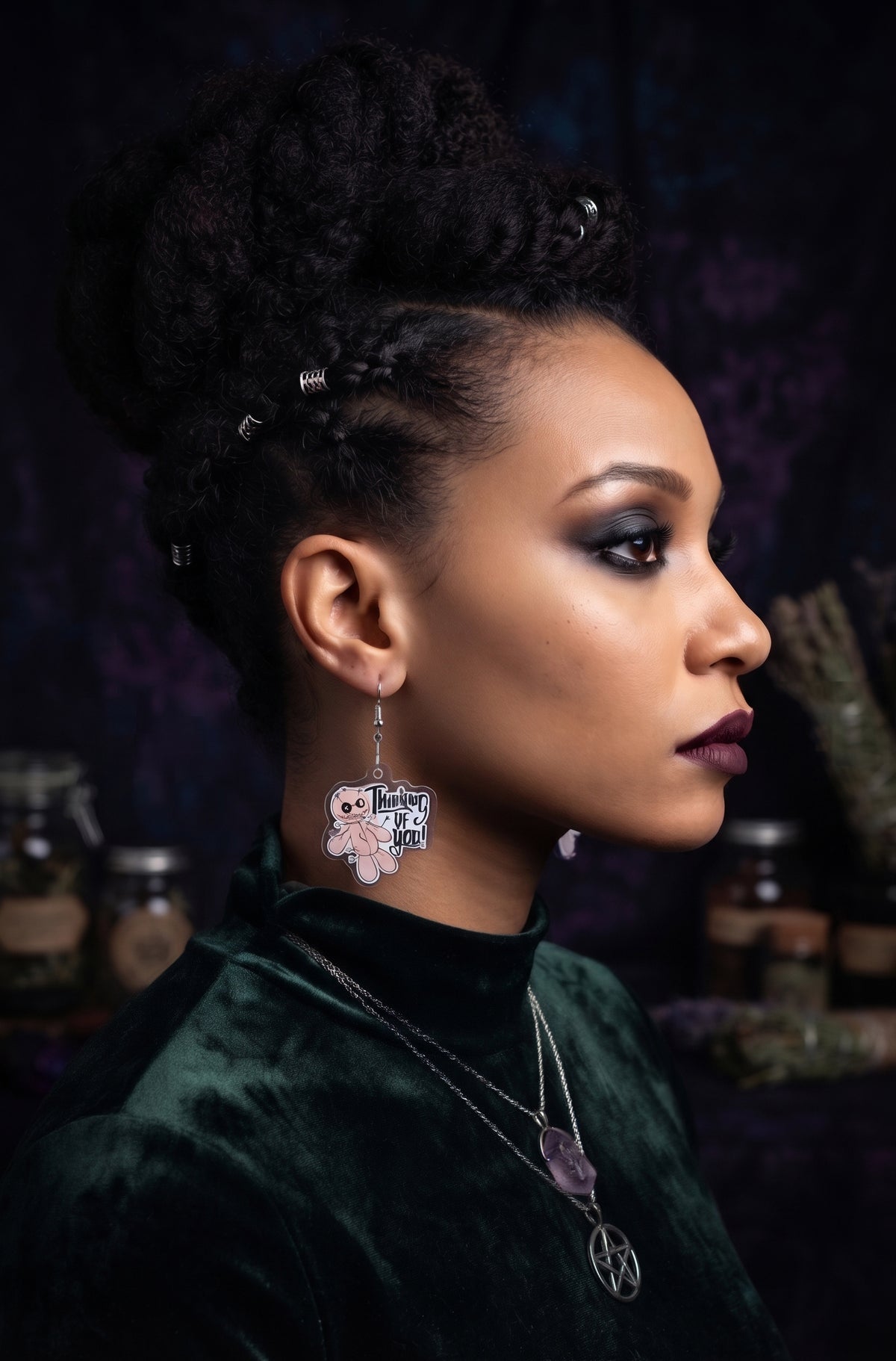 Voodoo Doll Thinking of You Earrings by Circle of Heather worn by a model with a curly updo and silver hair rings, printed acrylic voodoo doll charm reading "Thinking of you," shown with a green velvet top and pentacle necklace against apothecary shelves.
